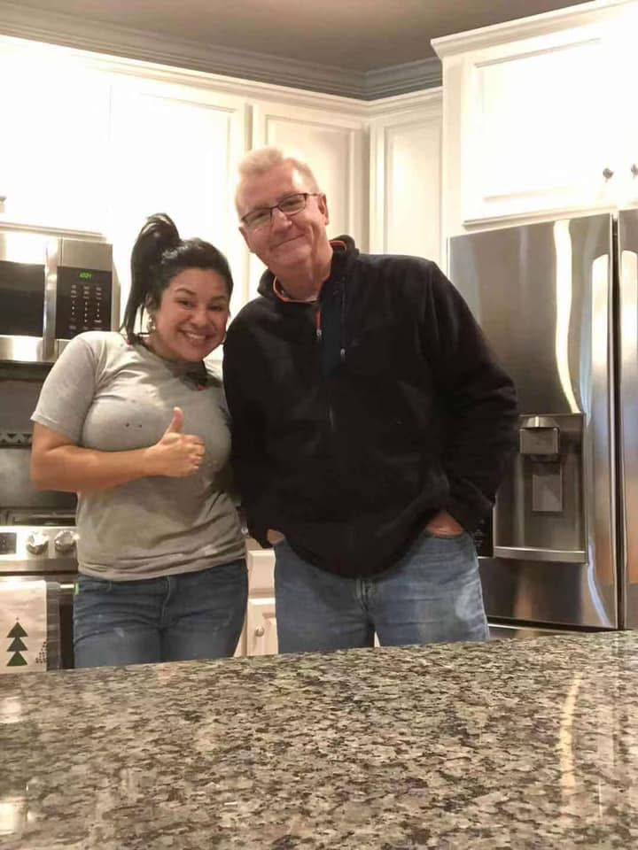 A woman giving a thumbs up and smiling stands next to a man with glasses who is also smiling. They are in a modern kitchen with white cabinets, a refrigerator, and granite countertops.