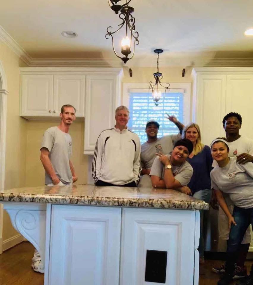 Six people pose and smile together in a bright kitchen, standing and leaning around a marble-topped island beneath two hanging lights. The atmosphere appears friendly and casual.