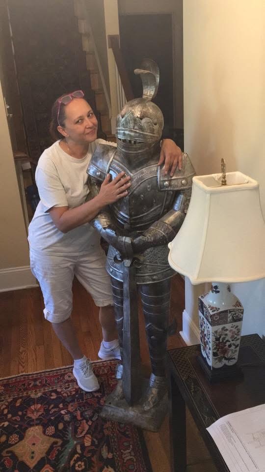A woman in white shorts and a t-shirt smiles while hugging a suit of medieval armor standing indoors next to a table with a lamp.