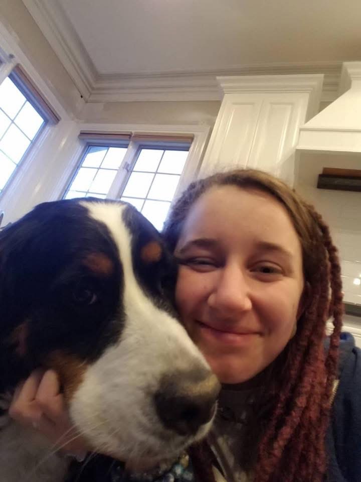 A person with long, braided hair smiles while posing closely with a large, black, brown, and white dog inside a bright kitchen with white cabinets and large windows.