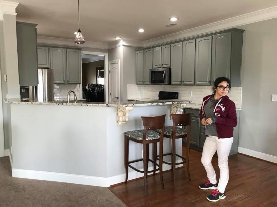 A woman wearing glasses, a burgundy jacket, and white pants stands near a kitchen counter with two wooden bar stools. The kitchen has gray cabinets, white backsplash, and stainless steel appliances.