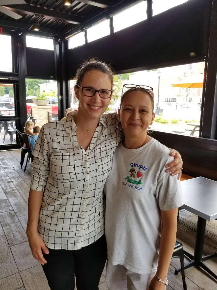 Two women stand closely together inside a cafe, smiling at the camera. The woman on the left wears glasses and a checkered shirt, while the woman on the right wears a graphic T-shirt. Tables and windows are visible in the background.
