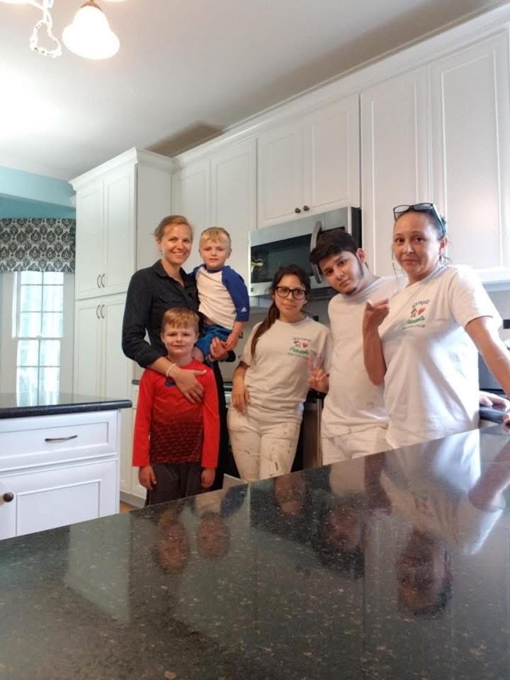 Five people, including two adults and three children, pose and smile together in a bright, modern kitchen with white cabinets and a dark countertop. The group appears relaxed and cheerful.