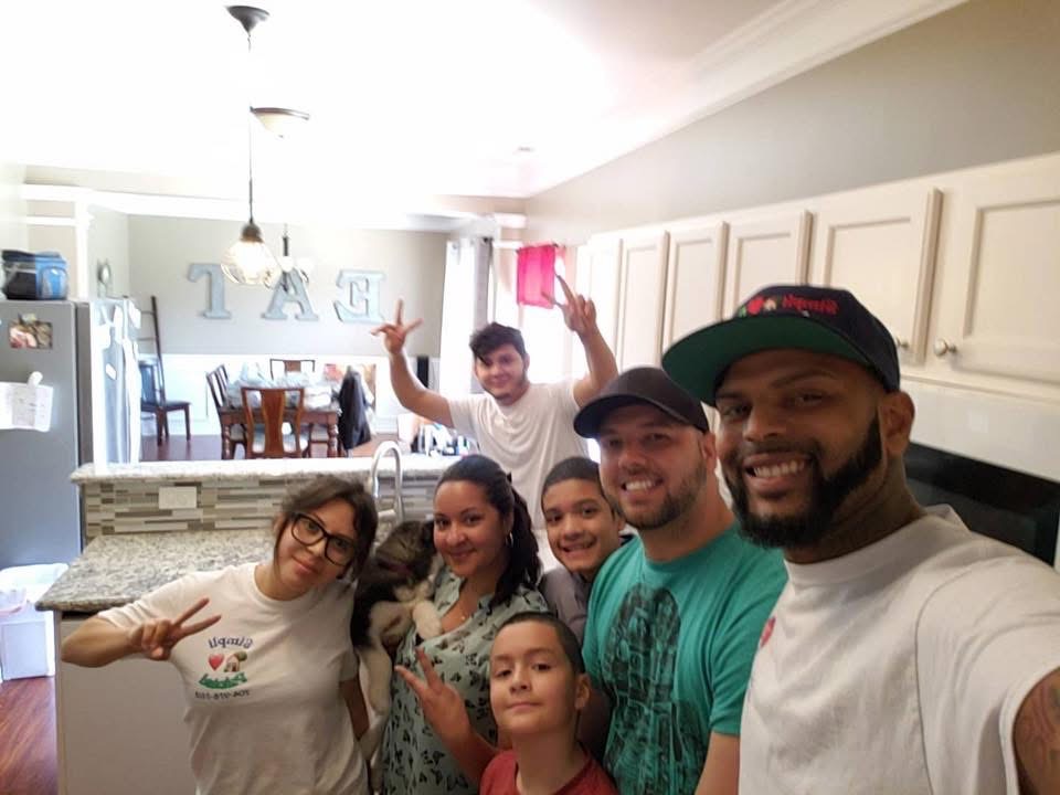 A group of six people, including two kids, pose and smile for a selfie in a bright kitchen. One person holds a small dog, and others make peace signs. The dining area and letters spelling EAT are visible in the background.