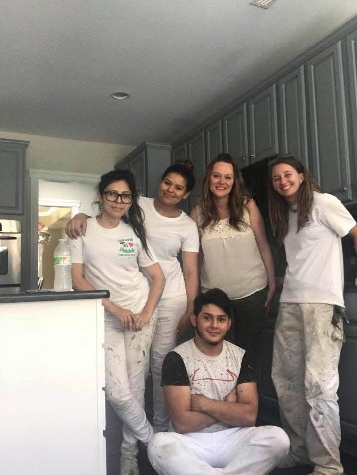 Five young adults, four women and one man, smile in a kitchen with grey cabinets. They are wearing paint-stained clothes, suggesting they are painting or renovating together. One woman is seated while the others stand behind her.