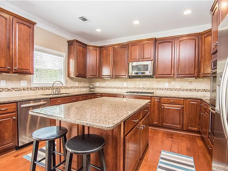 Spacious kitchen with wooden cabinets expertly finished by cabinet painters, granite island with two stools, stainless steel appliances, and tile backsplash. Large window above the sink provides natural light.