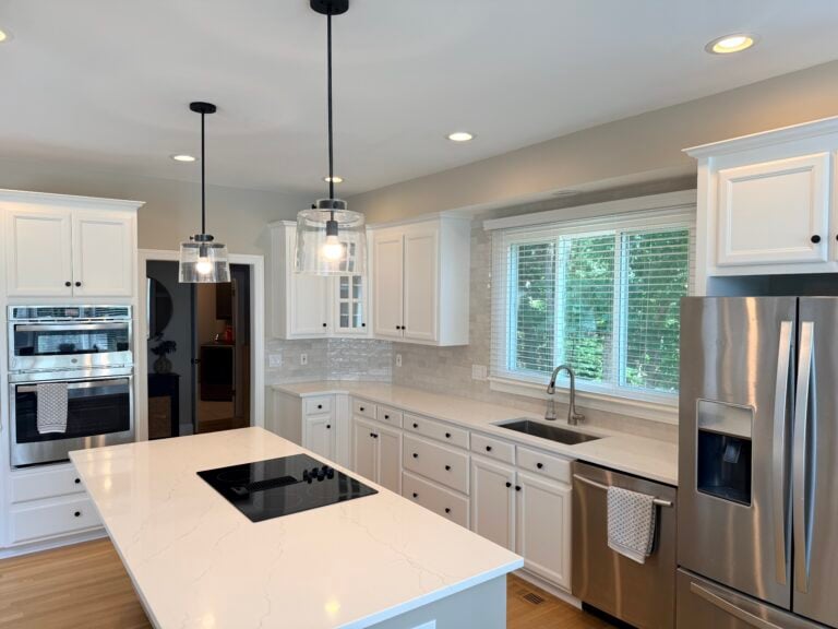 Modern kitchen with freshly painted white cabinets—offering a glimpse into cabinet painting cost—stainless steel appliances, an island with a stovetop, pendant lights, a large window above the sink, and light wood flooring.