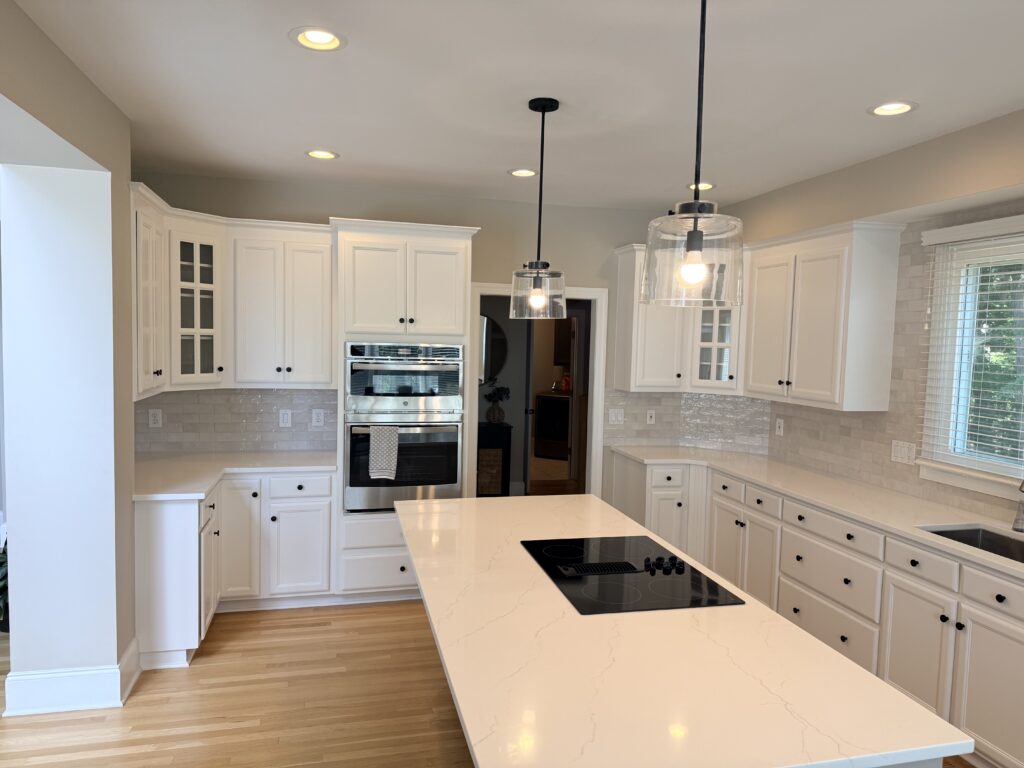Modern kitchen in Moorseville NC with white cabinets, a central island with a cooktop, double oven, pendant lights, light wood floors, and a large window letting in natural light.