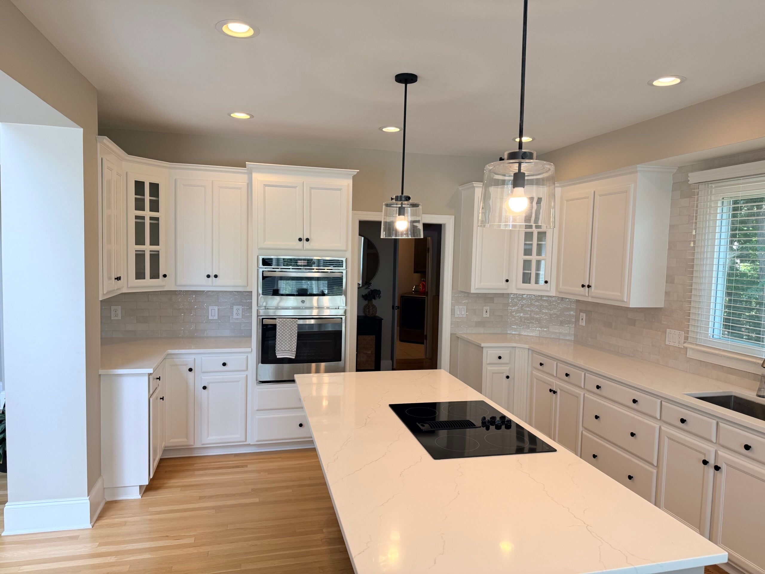 Modern kitchen in Moorseville NC with white cabinets, a central island with a cooktop, double oven, pendant lights, light wood floors, and a large window letting in natural light.