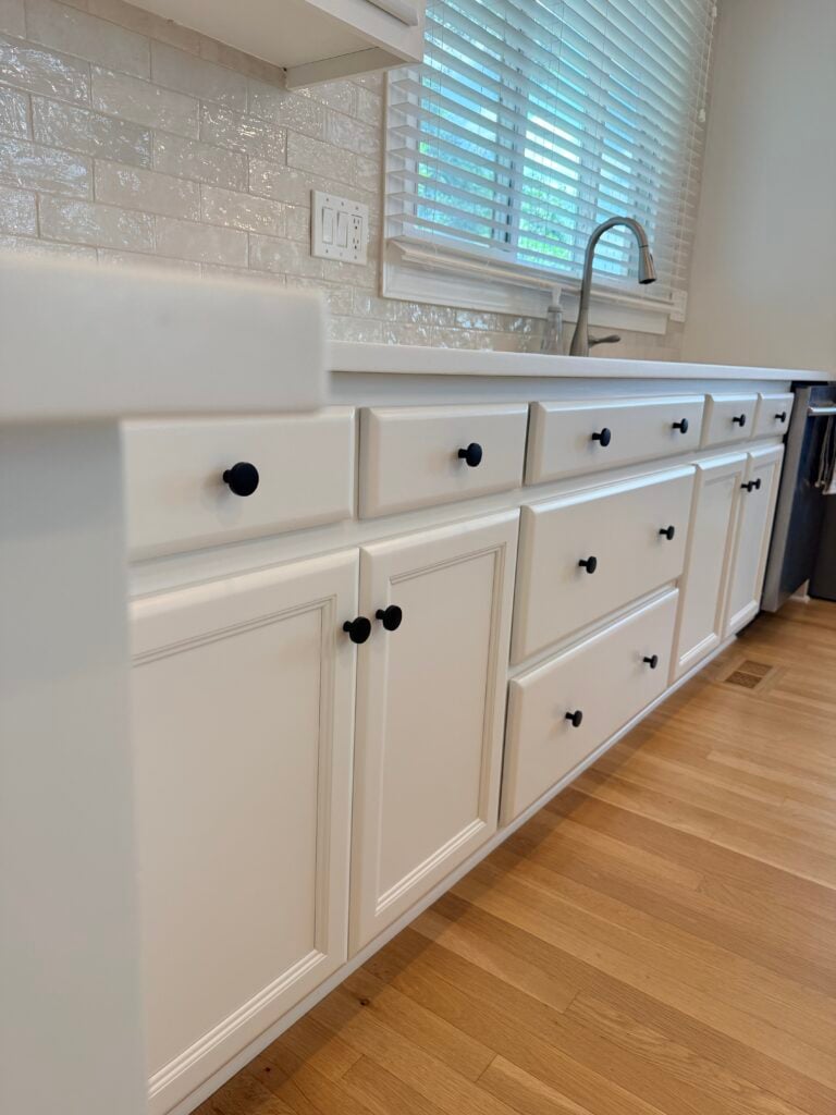 White kitchen cabinets with black knobs, under a white countertop. A sink with a silver faucet is visible, and there are wooden floors and a window with blinds above the countertop—creating a fresh look that highlights just how transformative a simpli painted upgrade can be without the high cabinet painting cost.