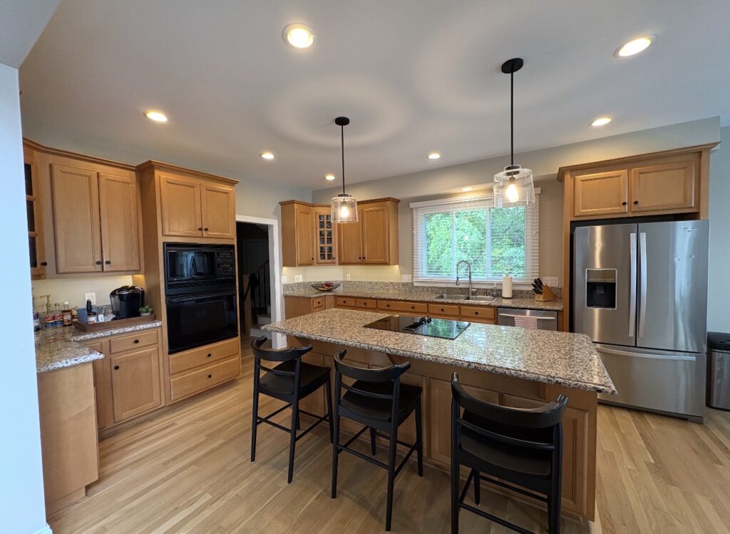 Spacious kitchen with freshly refinished wooden cabinets by expert cabinet painters, granite island with four chairs, stainless steel appliances, double oven, hanging lights, and a window above the sink showing greenery outside.