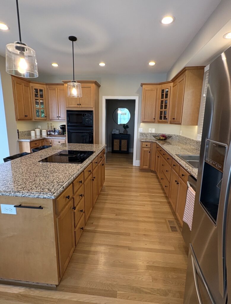 Spacious kitchen with wood cabinets expertly finished by professional cabinet painters, granite countertops, stainless steel appliances, and two pendant lights over a central island. Light wood floor and a doorway lead to another room.