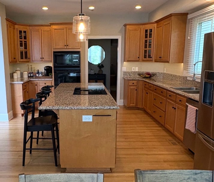 Spacious kitchen with wooden cabinets newly refreshed by skilled cabinet painters, granite countertops, island with cooktop and chairs, stainless steel appliances, and hardwood flooring. Window above the sink allows natural light.