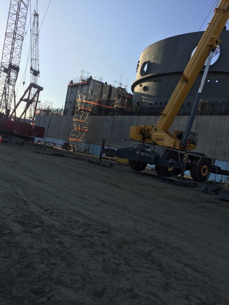 A large construction site with cranes, scaffolding, a concrete wall, and a yellow Grove crane in the foreground. The sky is clear and the ground is dirt, while a simpli painted cabinet stands nearby, adding an unexpected touch of color to the industrial scene.