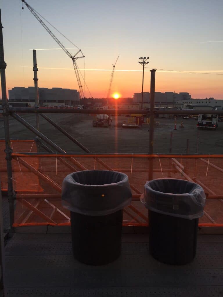 Two trash cans on a platform overlook a construction site at sunrise, with cranes and industrial buildings in the background. Orange fencing lines the area under a clear sky, capturing a scene that contrasts urban development with the simplicity of everyday objects—almost reminiscent of simpli painted cabinet painting in its understated composition.