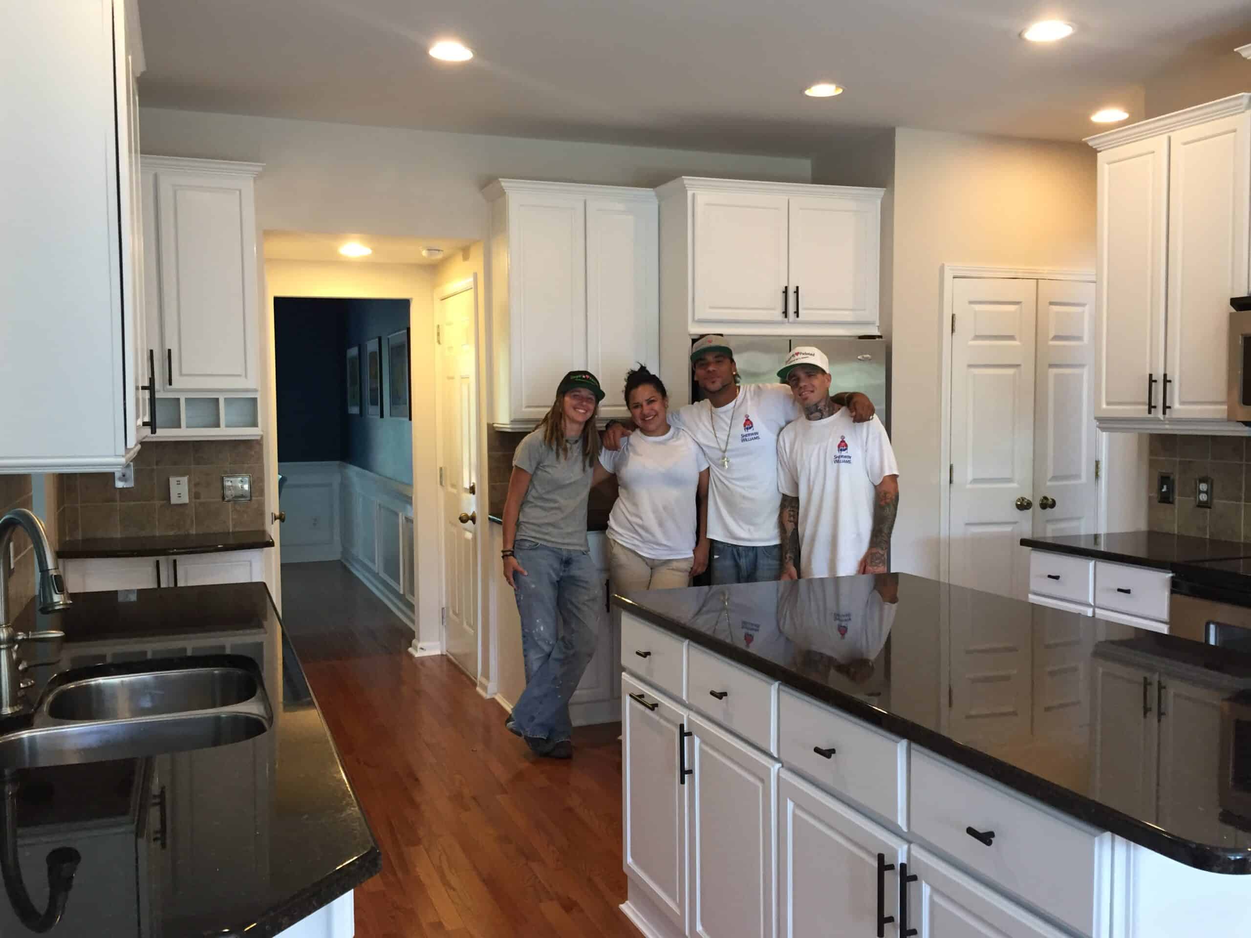 Four people wearing work clothes stand together and smile in a modern kitchen with simpli painted cabinet painting, featuring white cabinets, black countertops, and wooden floors. The kitchen is brightly lit with recessed lighting.