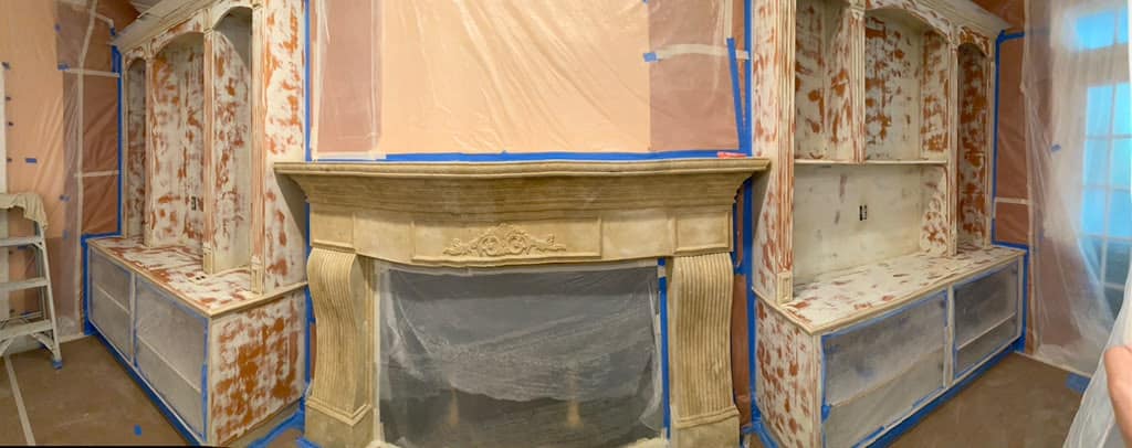 Wooden shelves and a fireplace are undergoing renovation. The walls, shelves, and fireplace are covered with protective plastic sheets and blue tape as part of the simpli painted cabinet painting process. Some areas show patches of exposed wood and primer.