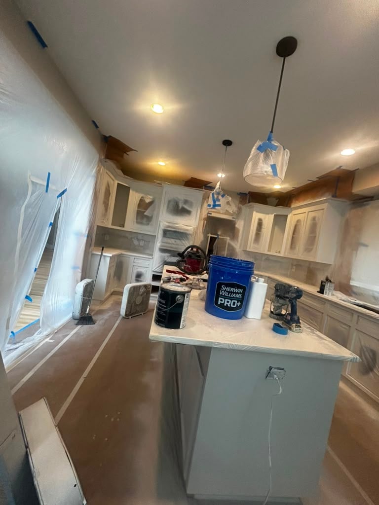 A kitchen under renovation with plastic sheets covering cabinets and appliances, supplies and tools on the island, and protective floor covering. Ceiling lights are on, blue painters tape is visible throughout the space, reflecting careful preparation for a Simpli Painted cabinet painting project.