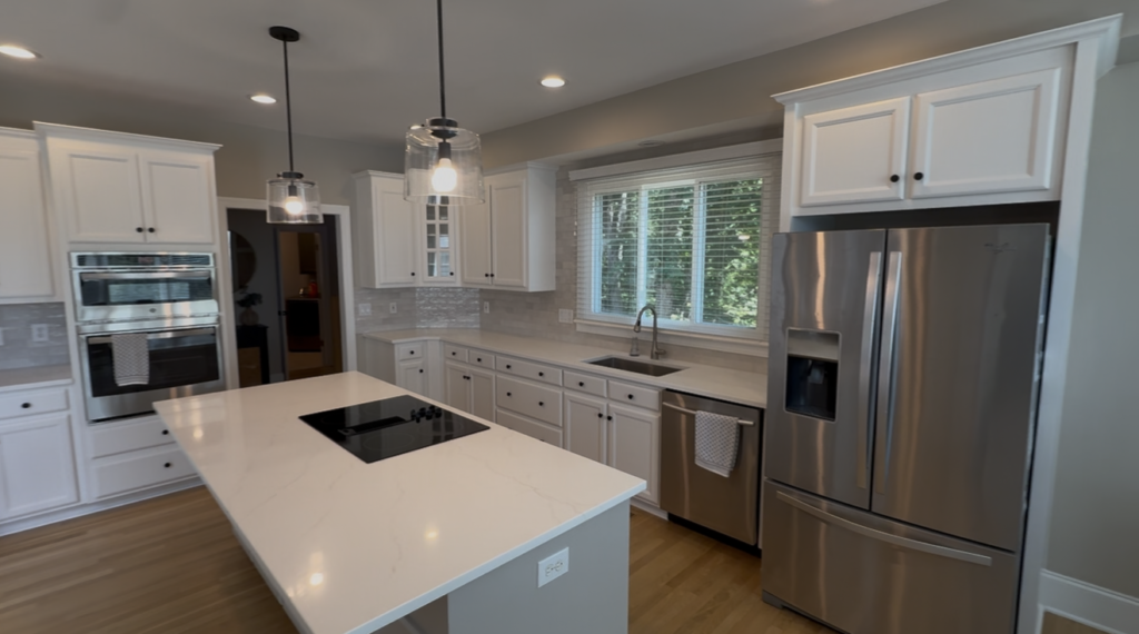 Modern kitchen with white cabinets expertly finished by skilled cabinet painters, stainless steel appliances, a large island with a cooktop, pendant lights, a double sink under a window, and wood flooring.