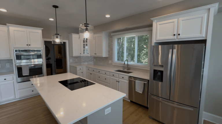 Modern kitchen with white cabinets expertly finished by skilled cabinet painters, stainless steel appliances, a large island with a cooktop, pendant lights, a double sink under a window, and wood flooring.
