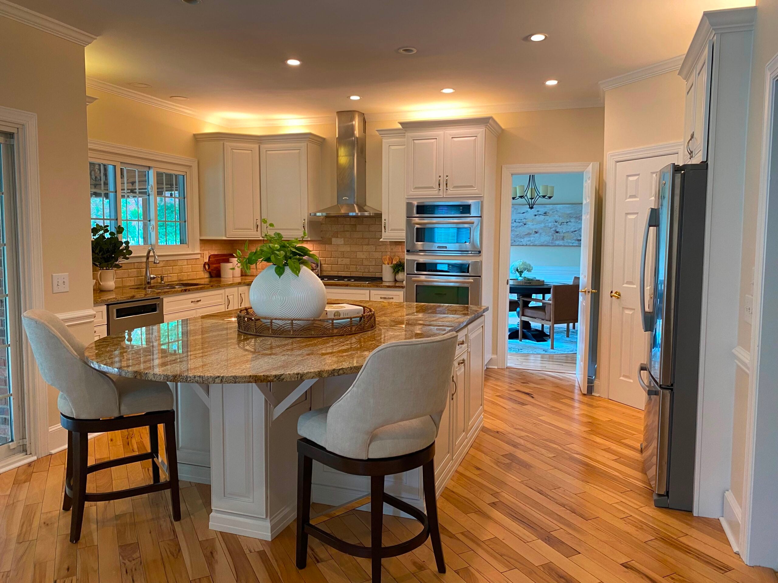 Modern kitchen with white cabinets, stainless steel appliances, a rounded granite island, two upholstered chairs, hardwood floors, and a view into a dining room through an open doorway.