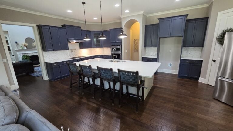 Modern kitchen with dark blue cabinets, large white island, four chairs, stainless steel fridge, pendant lights, and hardwood floors. Adjoining living area visible in the background.