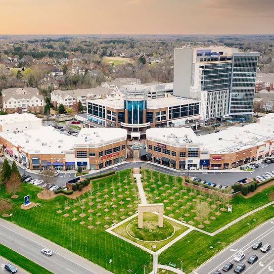 Aerial view of the modern Ballantyne business complex with landscaped green lawns, a central monument, parking lots, surrounding roads, and residential neighborhoods in the background under a cloudy sky.