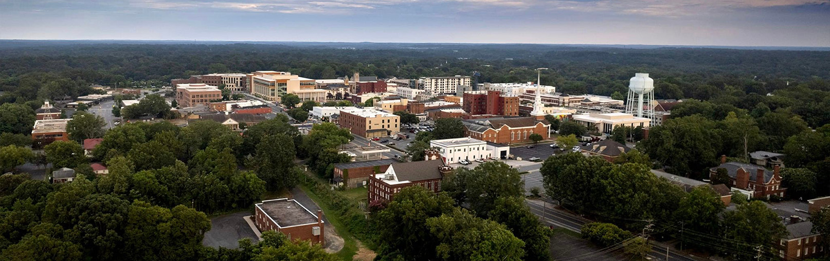 Aerial view of Concord, NC, a small city with a mix of trees, buildings, a water tower, and roads under a cloudy sky.
