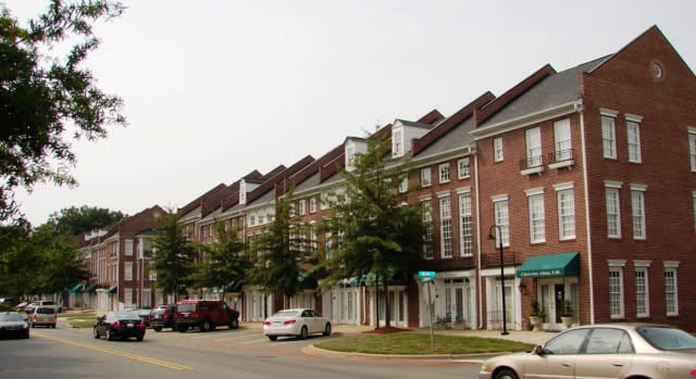 Cornelius NC. A row of brick townhouses with green awnings lines a quiet street in Cornelius, NC. Several cars are parked along the curb, and small trees are planted on the sidewalk in front of the buildings.