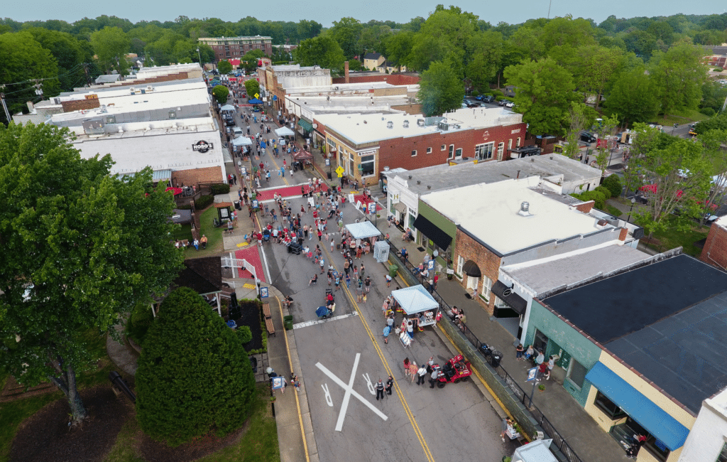 Aerial view of a small town street festival with crowds, vendor tents, and brick buildings—some recently refreshed by cabinet painting in Fort Mill professionals—surrounded by trees on a cloudy day.