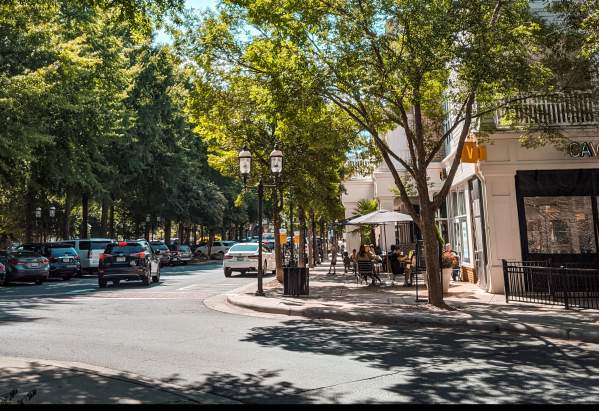 Huntersville NC. A sunny street in Huntersville, NC, lined with trees, parked cars, and people sitting at outdoor tables near a cafe. Buildings and storefronts are visible on the right side of the image.