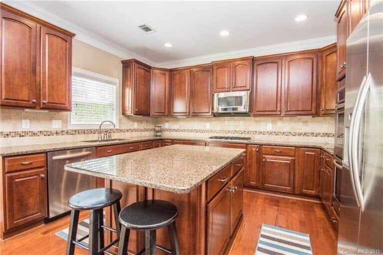 Spacious kitchen with wooden cabinets expertly finished by skilled cabinet painters, granite countertops, stainless steel appliances, an island with two black stools, and hardwood flooring. A window above the sink lets in natural light.