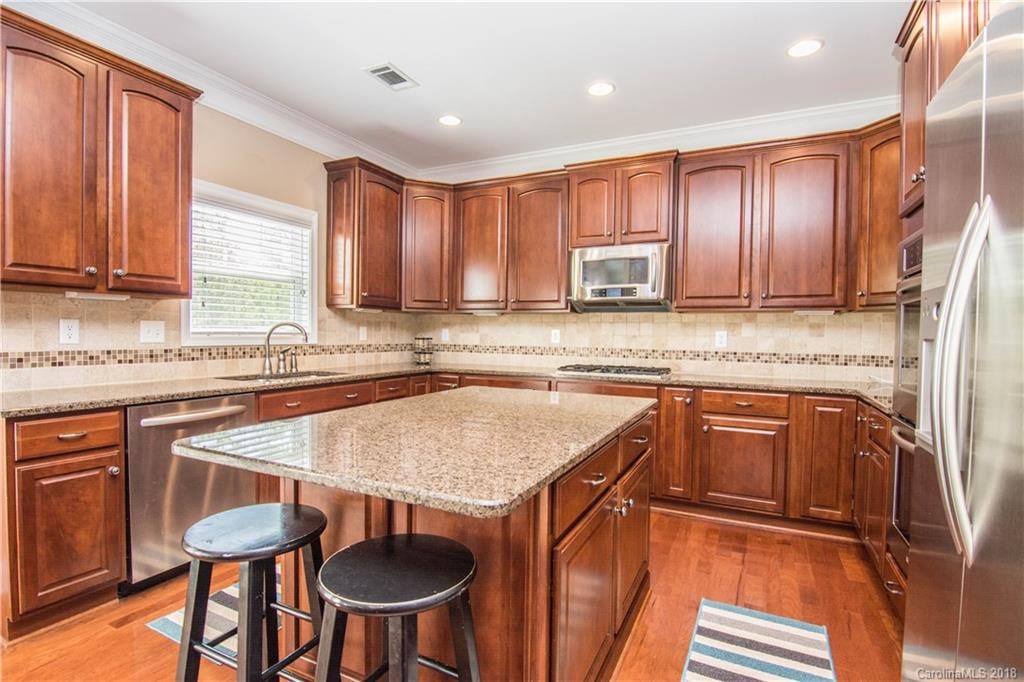 Spacious kitchen with professionally finished wooden cabinets by expert cabinet painters, granite countertops, an island with two black stools, stainless steel appliances, and a window above the sink.