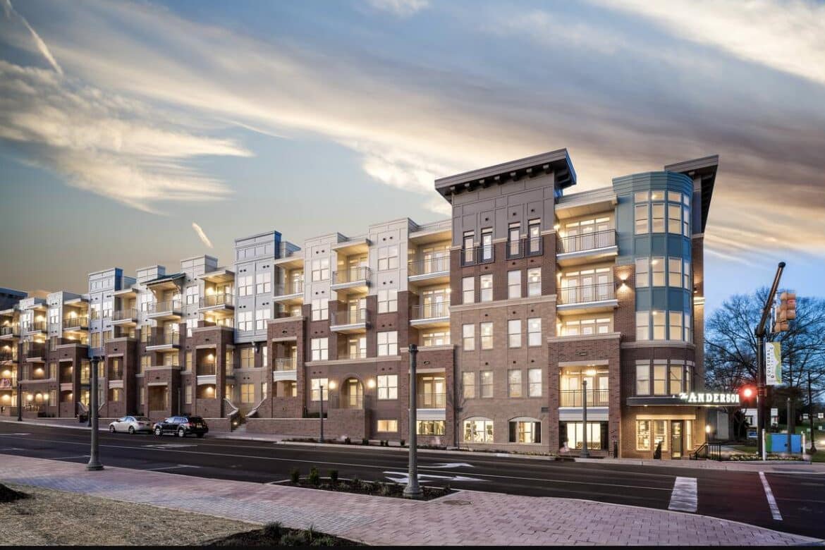 Modern multi-story apartment building with large windows, balconies, and illuminated interiors at dusk in Rock Hill, SC. Cars are parked along the street, and trees are visible in the background.