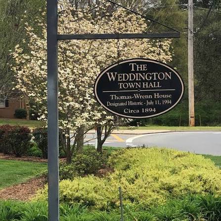 An oval sign reads: The Weddington Town Hall, Thomas-Wrenn House, Designated Historic July 11, 1994, Circa 1894. Shrubs, a tree, and a road are visible in the background—capturing a charming local scene that reflects the timeless character often celebrated by cabinet painting - Weddington NC.