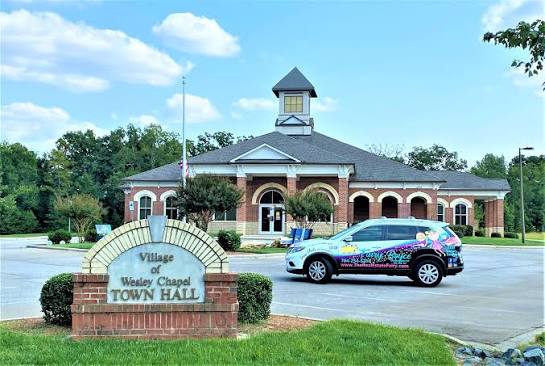 A brick town hall building with a clock tower stands behind a sign reading Village of Wesley Chapel Town Hall. A colorful van advertising cabinet refinishing in Wesley Chapel is parked in front. Trees and a blue sky are in the background.